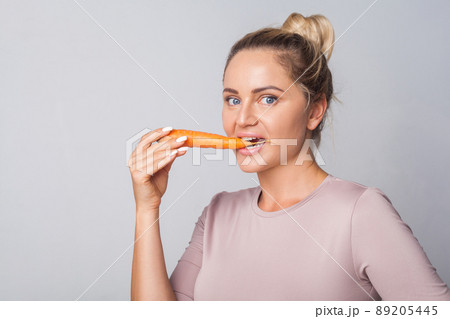 Portrait of young woman biting orange carrot, fresh raw vegetables, concept of healthy eating with vitamins beta carotene, vegetarian diet, low calorie food. studio shot isolated on grey background 89205445