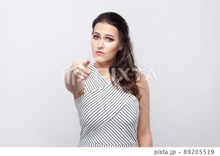 You. Portrait of serious beautiful young brunette woman with makeup and striped dress standing, looking and pointing at camera. indoor studio shot, isolated on grey background. You. Portrait of serious beautiful young brunette woman with makeup and striped dress standing, looking and pointing at camera. indoor studio shot, isolated on grey background. 89205519