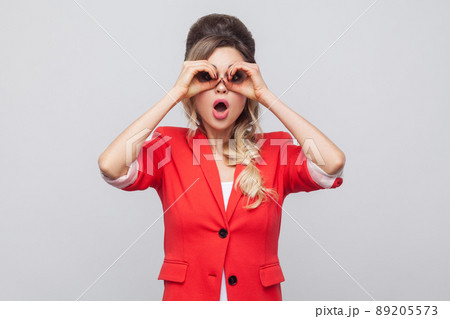 Portrait of surprised beautiful business lady with hairstyle and makeup in red fancy blazer, standing and looking in binoculars gesture. indoor studio shot, isolated on grey background. Portrait of surprised beautiful business lady with hairstyle and makeup in red fancy blazer, standing and looking in binoculars gesture. indoor studio shot, isolated on grey background. 89205573