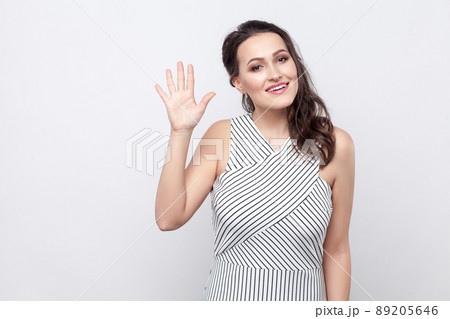 Portrait of beautiful happy young brunette woman with makeup and striped dress standing, looking at camera with toothy smile and greeting gesture. indoor studio shot, isolated on grey background. Portrait of beautiful happy young brunette woman with makeup and striped dress standing, looking at camera with toothy smile and greeting gesture. indoor studio shot, isolated on grey background. 89205646