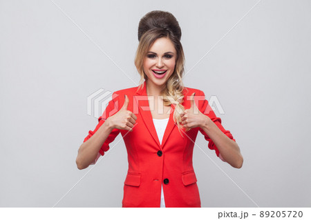 Portrait of glad beautiful business lady with hairstyle and makeup in red fancy blazer, standing, thumbs up and looking at camera with thumbs up. indoor studio shot, isolated on grey background. 89205720
