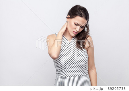 Neck or back pain. Portrait of sick young brunette woman with makeup and striped dress standing holding her neck and feeling pain. indoor studio shot, isolated on grey background. 89205733