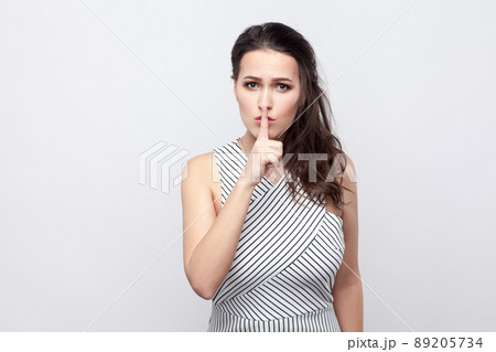 Portrait of serious beautiful young brunette woman with makeup and striped dress standing and looking at camera with warning sign. indoor studio shot, isolated on grey background. 89205734