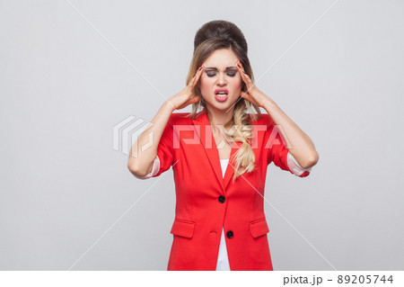 Portrait of sick beautiful business lady with hairstyle and makeup in red fancy blazer standing, holding her head and feeling headache. indoor studio shot, isolated on grey background. 89205744