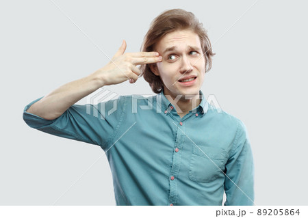 Portrait of funny or sad handsome long haired blonde young man in blue casual shirt standing with gun pistol gesture hand on his head. indoor studio shot, isolated on light grey background. 89205864