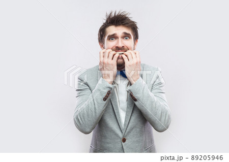 Portrait of worry handsome bearded man in casual grey suit and blue bow tie standing, nervous bitting his nails and looking at camera. indoor studio shot, isolated on light grey background. 89205946