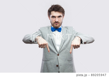 Here and right now. Portrait of serious bossy handsome bearded man in casual grey suit and blue bow tie standing pointing and looking at camera. indoor studio shot, isolated on light grey background. 89205947