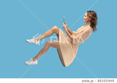 Hovering in air. Smiling girl in yellow dress levitating with mobile phone, reading message chatting happy joyful in social network online, surfing web while flying. indoor shot isolated on blue Hovering in air. Smiling girl in yellow dress levitating with mobile phone, reading message chatting happy joyful in social network online, surfing web while flying. indoor shot isolated on blue 89205949
