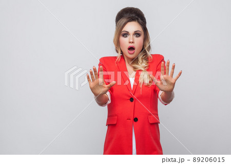 Portrait of shocked beautiful business lady with hairstyle and makeup in red fancy blazer, standing with blocking hands and looking at camera. indoor studio shot, isolated on grey background. 89206015