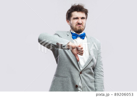 Dislike. Portrait of dissatisfied handsome bearded man in casual grey suit and blue bow tie standing and looking at camera with thumbs down. indoor studio shot, isolated on light grey background. Dislike. Portrait of dissatisfied handsome bearded man in casual grey suit and blue bow tie standing and looking at camera with thumbs down. indoor studio shot, isolated on light grey background. 89206053