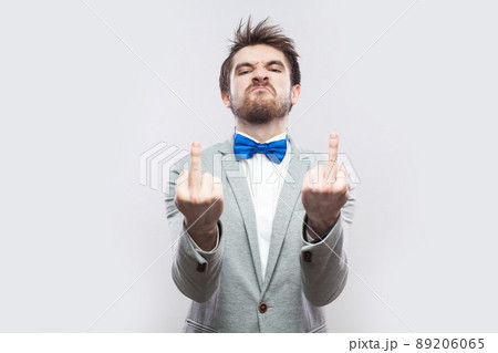Portrait of angry handsome bearded man in casual grey suit and blue bow tie standing with middle finger fuck sing and looking at camera. indoor studio shot, isolated on light grey background. Portrait of angry handsome bearded man in casual grey suit and blue bow tie standing with middle finger fuck sing and looking at camera. indoor studio shot, isolated on light grey background. 89206065