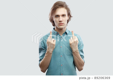 Portrait of poker face handsome long haired blonde young man in blue casual shirt standing and looking at camera with middle finger fuck sign. indoor studio shot, isolated on light grey background. 89206369