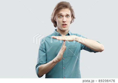 I need more time. Portrait of worry handsome long haired blonde young man in blue casual shirt standing with time out sign and looking at camera. indoor studio shot, isolated on light grey background. 89206476