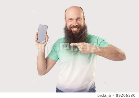 Portrait of happy satisfied middle aged bald bearded man standing with mobile smart phone pointing at empty screen and looking with toothy smile. indoor studio shot, isolated on grey background. 89206530