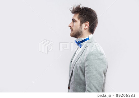 Profile side view portrait of serious handsome bearded man in casual grey suit, blue bow tie standing and looking straight wih calm serious face. indoor studio shot, isolated on light grey background. 89206533