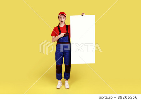 Full length portrait of astonished worker woman holding and pointing at white paper placard with empty space for advertisement or promotional text. Indoor studio shot isolated on yellow background. 89206556