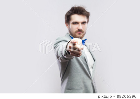 Portrait of serious handsome bearded man in casual grey suit and blue bow tie standing and looking and blaming at camera. indoor studio shot, isolated on light grey background. Portrait of serious handsome bearded man in casual grey suit and blue bow tie standing and looking and blaming at camera. indoor studio shot, isolated on light grey background. 89206596