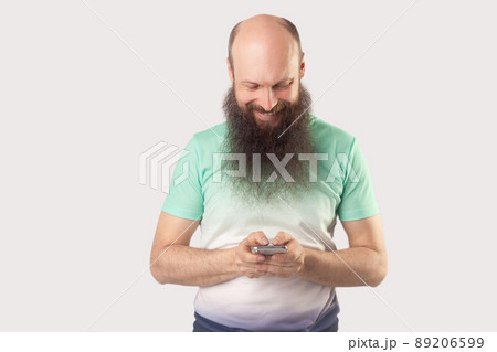 Portrait of happy middle aged bald bearded man standing, holding mobile smart phone and looking at screen, reading something with toothy smile. indoor studio shot, isolated on grey background. Portrait of happy middle aged bald bearded man standing, holding mobile smart phone and looking at screen, reading something with toothy smile. indoor studio shot, isolated on grey background. 89206599