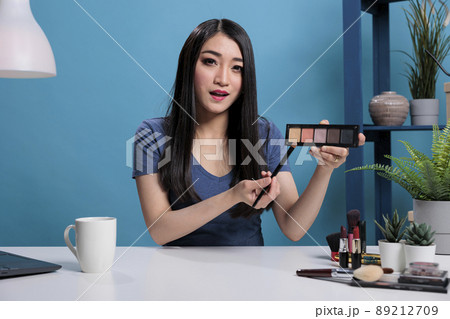 Asian woman influencer holding palette showing at camera while doing makeup tutorial advertising beauty product. Social media content creator recording cosmetic review for vlogging channel 89212709