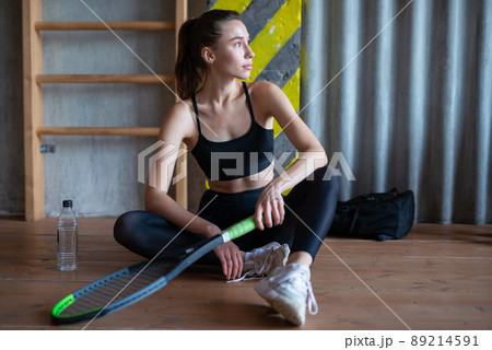 Portrait of female tennis player with racket sitting on gym floor Portrait of female tennis player with racket sitting on gym floor 89214591