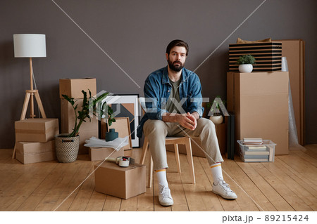 Horizontal shot portrait of young Caucasian man with beard on face sitting in loft room with stuff packed for moving to new apartment 89215424