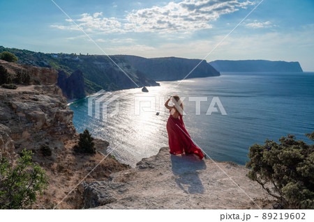 A girl with flowing hair in a long red dress stands on a rock above the sea. The stone can be seen in the sea. Sunny path to the sea from the sun. 89219602