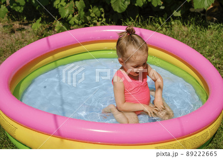 A cute little girl in a swimsuit splashes in the water in a small multicolored inflatable swimming pool. A hot sunny summer day. A cute little girl in a swimsuit splashes in the water in a small multicolored inflatable swimming pool. A hot sunny summer day. 89222665