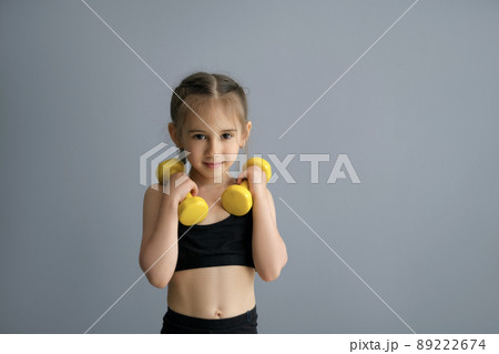 Cute beautiful sporty girl with braided pigtails plays fitness by lifting heavy dumbbells. Healthy, strong child in a sportswear . Person with sports equipment on a grey studio background copy space Cute beautiful sporty girl with braided pigtails plays fitness by lifting heavy dumbbells. Healthy, strong child in a sportswear . Person with sports equipment on a grey studio background copy space 89222674