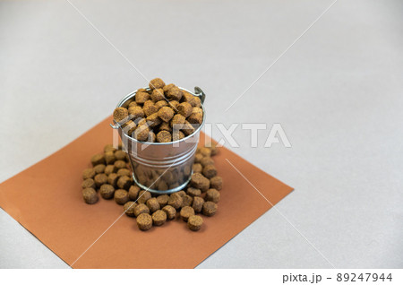 A full metal bucket of dry pelleted dog food. A small silver thigh of dog food stands against a gray background. A square brown piece of paper with a scattering of pellets. A full metal bucket of dry pelleted dog food. A small silver thigh of dog food stands against a gray background. A square brown piece of paper with a scattering of pellets. 89247944