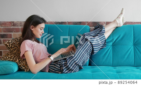 A young freelance woman in home clothes, working remotely in a home office, uses a laptop while lying on the couch. A modern woman browses the Internet in search of information, chatting online with 89254064