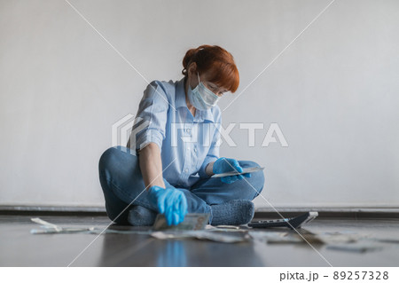 An adult woman in a medical mask on her face sits on the floor and counts money on a calculator, on a white background 89257328
