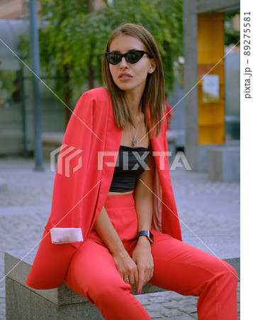 Blonde girl in a red lady-type pantsuit and black top, watch, ring, pendant and sunglasses is posing sitting on a stone bench. Close-up shot. 89275581