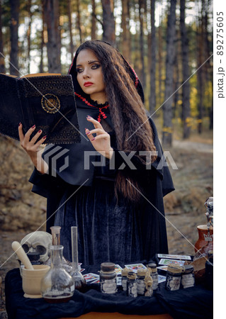 Witch in black dress with cape and hood. Holding book, pronouncing spell, posing in pine forest at table with accessories for witchcraft. Close-up. Witch in black dress with cape and hood. Holding book, pronouncing spell, posing in pine forest at table with accessories for witchcraft. Close-up. 89275605