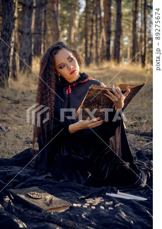Witch in black dress with cape and hood. Posing in pine forest. Sitting on dark blanket whith books, candles and runes on it. Full length. Witch in black dress with cape and hood. Posing in pine forest. Sitting on dark blanket whith books, candles and runes on it. Full length. 89275674