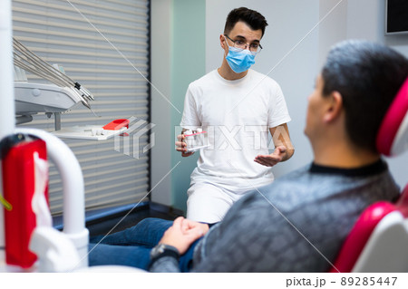 Dental surgeon with dental crown in hands model talks to patient about prosthetics. Man at the dentist's appointment Dental surgeon with dental crown in hands model talks to patient about prosthetics. Man at the dentist's appointment 89285447