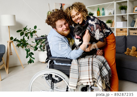 Young bearded man in wheelchair spending rehabilitation at home together with his wife and lovely dog Young bearded man in wheelchair spending rehabilitation at home together with his wife and lovely dog 89287235