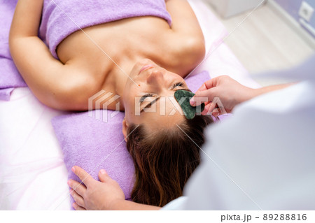Relaxing massage. European woman getting quartz guache face massage in spa salon, side view Relaxing massage. European woman getting quartz guache face massage in spa salon, side view 89288816