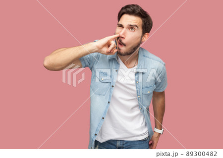 Portrait of crazy handsome bearded young man in blue casual style shirt standing and drilling his nose with funny face. indoor studio shot, isolated on pink background. Portrait of crazy handsome bearded young man in blue casual style shirt standing and drilling his nose with funny face. indoor studio shot, isolated on pink background. 89300482
