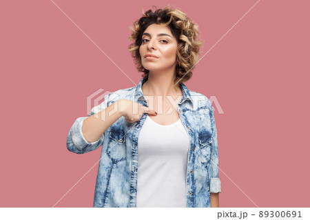 This is me. Portrait of serious young woman with curly hairstyle in casual blue shirt standing, pointing herself with proud face and looking at camera. indoor studio shot, isolated on pink background. This is me. Portrait of serious young woman with curly hairstyle in casual blue shirt standing, pointing herself with proud face and looking at camera. indoor studio shot, isolated on pink background. 89300691