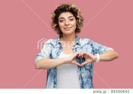 Portrait of happy young woman with curly hairstyle in casual blue shirt standing with heart hands shape gesture and looking at camera smiling. indoor studio shot, isolated on pink background. 89300692