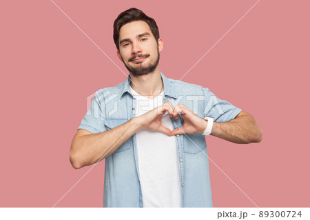 Portrait of happy smiley handsome bearded young man in blue casual style shirt standing with love hand gesture on heart and looking at camera smiling. indoor studio shot, isolated on pink background. Portrait of happy smiley handsome bearded young man in blue casual style shirt standing with love hand gesture on heart and looking at camera smiling. indoor studio shot, isolated on pink background. 89300724
