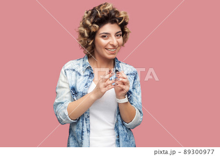 Portrait of funny cunning young woman with curly hairstyle in casual blue shirt standing and looking at camera with toothy smile playful face. indoor studio shot, isolated on pink background. 89300977