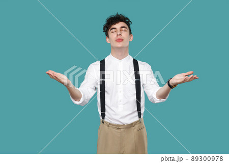 happy in love man raised arms and sending air kiss. Portrait of handsome hipster curly young businessman in classic casual white shirt and suspender standing. studio shot isolated on blue background. happy in love man raised arms and sending air kiss. Portrait of handsome hipster curly young businessman in classic casual white shirt and suspender standing. studio shot isolated on blue background. 89300978