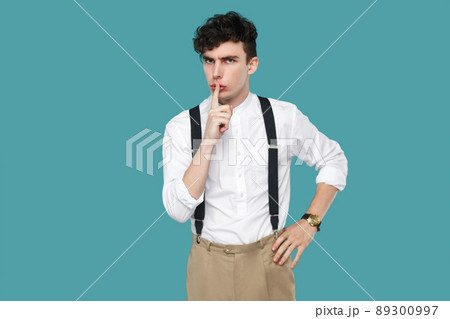Serious man showing silent sign and looking at camera. Portrait of handsome hipster curly young businessman in classic casual white shirt, suspender standing. studio shot isolated on blue background. 89300997