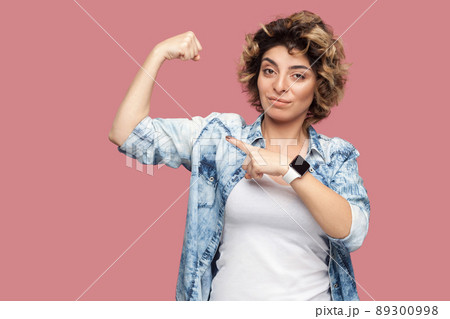 Portrait of strong proud young woman with curly hairstyle in casual blue shirt standing and pointing at her biceps to show her independent or feminism. indoor studio shot, isolated on pink background. 89300998