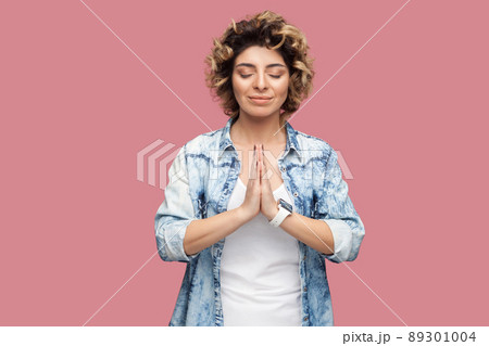 Portrait of calm young woman with curly hairstyle in casual blue shirt standing with closed eyes, palm hands, smile and doing yoga meditating. indoor studio shot, isolated on pink background. Portrait of calm young woman with curly hairstyle in casual blue shirt standing with closed eyes, palm hands, smile and doing yoga meditating. indoor studio shot, isolated on pink background. 89301004