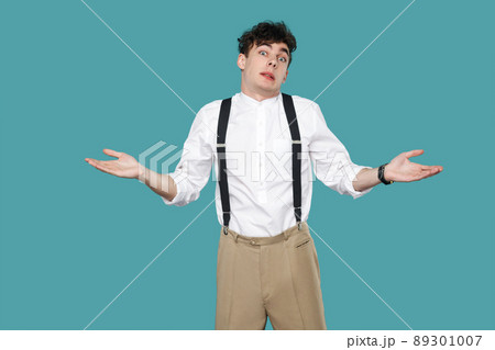 I don't know. confused man raised arms and looking at camera. Portrait of handsome hipster curly young businessman in classic casual white shirt and suspender. studio shot isolated on blue background. 89301007