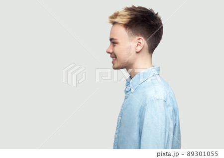 Profile side view portrait of happy handsome young man in light blue shirt standing with toothy smile and looking forward with happiness. indoor studio shot on grey background copy space. 89301055