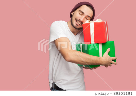 Portrait of happy satisfied handsome bearded young hipster man in white shirt and casual hat standing and holding two present bo and hugging it. Indoor, isolated, studio shot, pink background 89301191