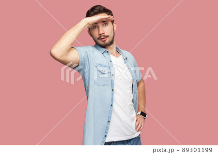 Portrait of attentive handsome bearded young man in blue casual style shirt standing with hand on forehead and looking away. indoor studio shot, isolated on pink background. Portrait of attentive handsome bearded young man in blue casual style shirt standing with hand on forehead and looking away. indoor studio shot, isolated on pink background. 89301199